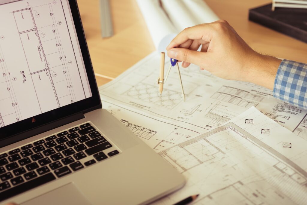 Architect working on blueprint in workplace with laptop and drawing compass.